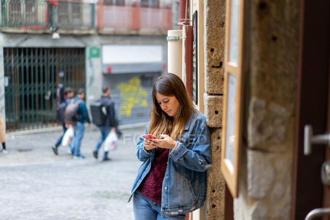 Attraktive Frau in Jeansjacke hält rotes Handy in der Hand und steht neben einer Mauer auf der Straße in Porto, Portugal — Stockfoto