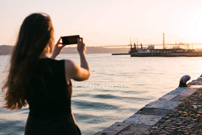 Вид женщины, стреляющей лодкой на смартфоне на набережной возле воды на закате — стоковое фото