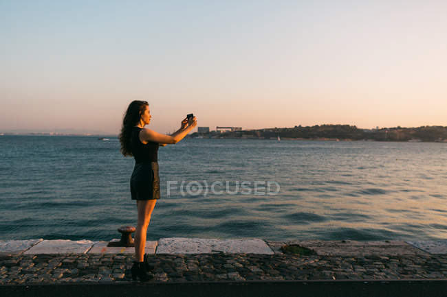 Giovane donna in abito nero scattare foto con smartphone su argine vicino all'acqua al tramonto — Foto stock