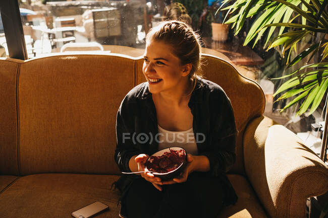 Hübsche junge Frau mit Schüssel und Löffel lächelt, während sie auf einer bequemen Couch am Fenster sitzt — Stockfoto