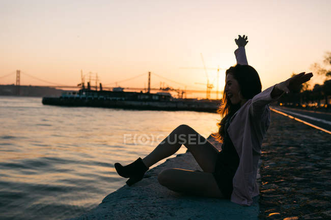 Весела молода жінка сидить на набережній біля поверхні води з кораблем на заході сонця — стокове фото