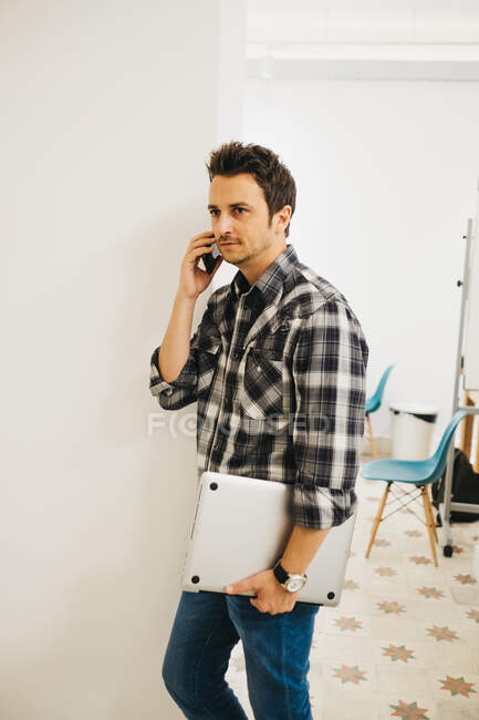Hombre joven con el ordenador portátil que habla en el teléfono inteligente - foto de stock