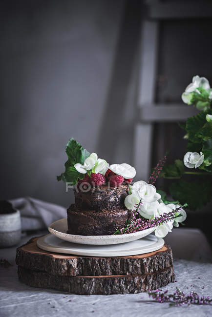 Chocolate cake decorated with raspberries and flowers served on plate on wooden stand — Stock Photo