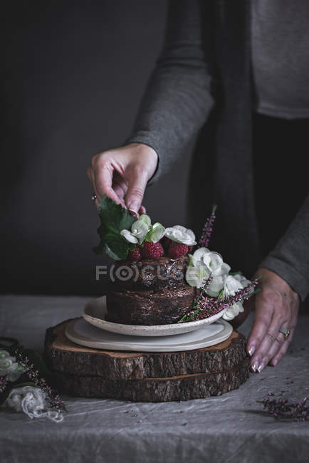 Female hands decorating chocolate cake decorated with raspberries and flowers on plate on wooden stand — Stock Photo