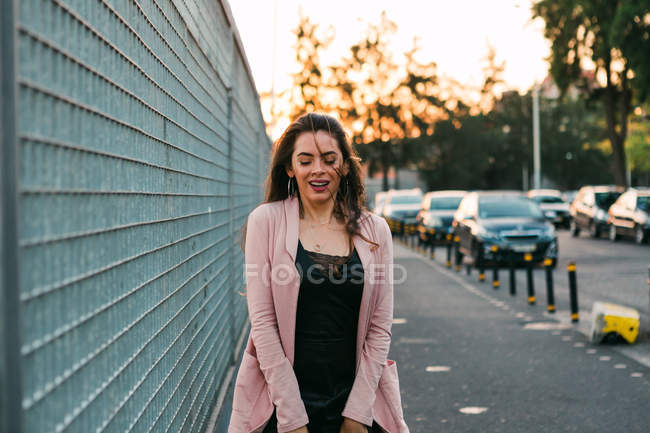 Усміхнена молода жінка, що йде вулицею біля автомобілів на заході сонця — стокове фото
