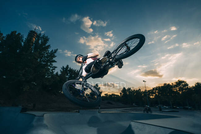 Giovane sportivo che fa trucchi su BMX in parco sullo sfondo del cielo nuvoloso blu — Foto stock