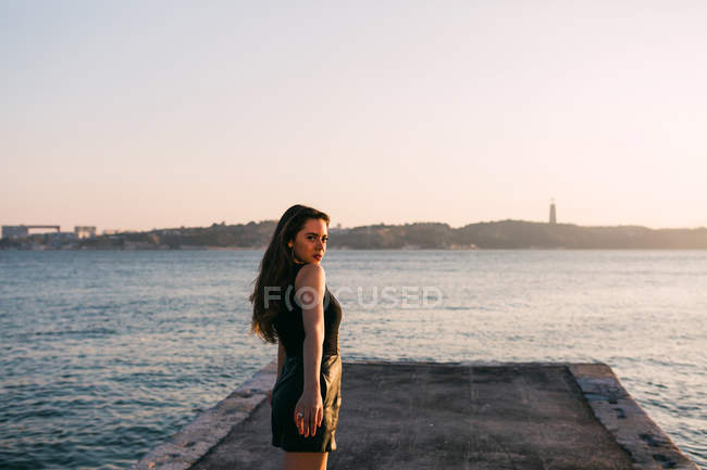 Спокуслива молода жінка в чорному платті, що стоїть на набережній біля поверхні води на заході сонця — стокове фото