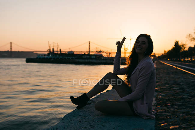 Lächelnde junge Frau sitzt auf einem Damm nahe der Wasseroberfläche mit Schiff bei Sonnenuntergang und zeigt ok Zeichen — Stockfoto