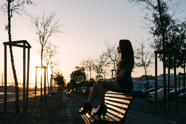 Sensual joven en vestido negro sentado en el banco en la calle en el callejón al atardecer - foto de stock