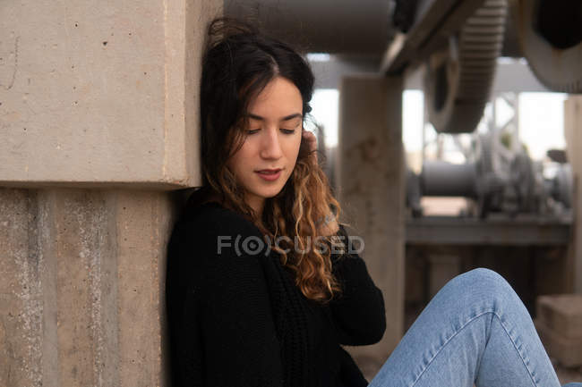 Encantadora jovem com cabelo encaracolado sentado perto da parede do edifício de concreto — Fotografia de Stock