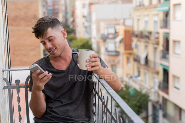 Расслабляющий мужчина пьет кофе и смотрит телефон на балконе — стоковое фото
