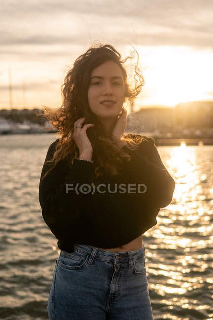 Adorável jovem mulher olhando para a câmera enquanto estava perto de água no aterro da cidade durante o pôr do sol — Fotografia de Stock