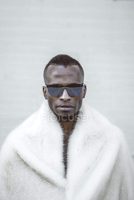 Handsome African American guy in trendy fur coat and sunglasses standing near white wall — Stock Photo
