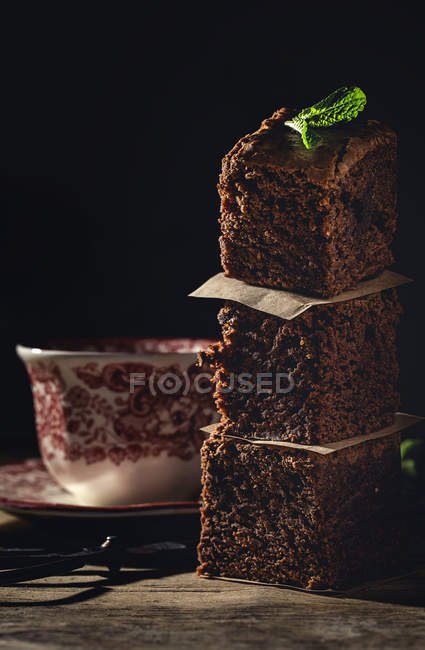 Stacked pieces of chocolate brownie with mint on dark background with cup of coffee — Stock Photo