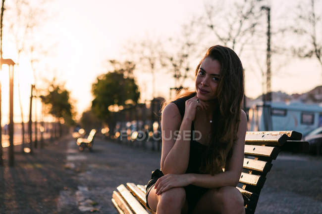 Retrato de jovem mulher de vestido preto sentado no banco na rua ao pôr do sol — Fotografia de Stock