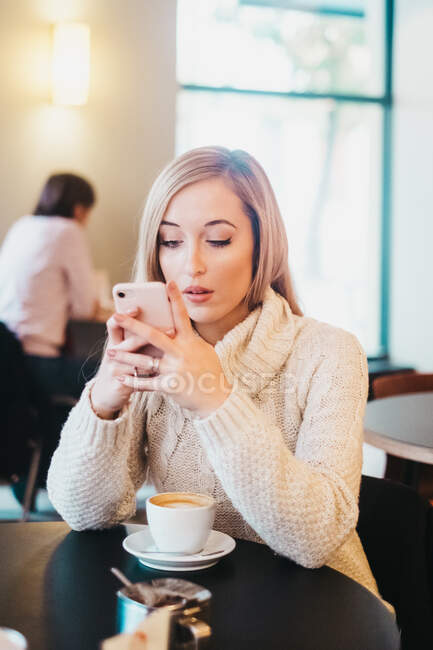 Jovem senhora atraente em suéter segurando telefone celular e sentado à mesa perto de caneca de café no café — Fotografia de Stock