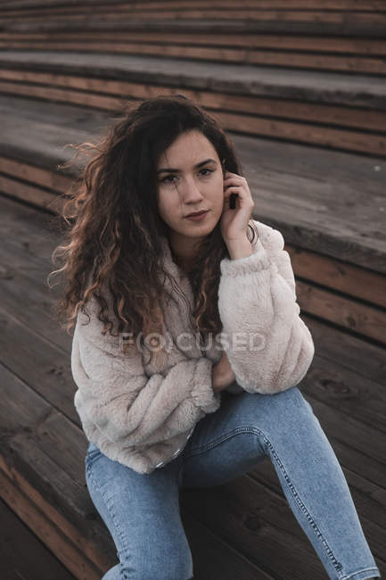 Attractive young woman in stylish outfit looking at camera while sitting on wooden stairs on city street — Stock Photo