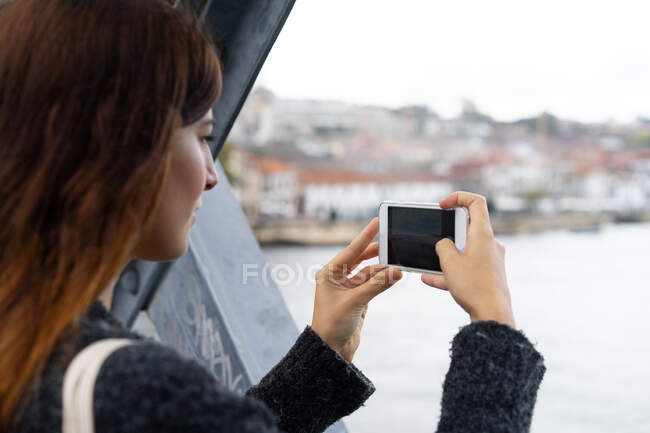 Eine junge Frau hält ihr Handy in der Hand und schießt von einer Brücke in Porto, Portugal, Wasser zwischen die Stadt — Stockfoto