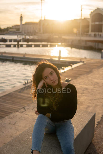 Jeune femme réfléchie regardant la caméra tout en étant assis près de l'eau sur le remblai de la ville pendant le coucher du soleil — Photo de stock