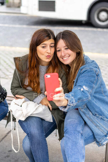Filles joyeux attrayant en tenue décontractée prendre selfie sur téléphone portable rouge et assis sur le siège dans la rue à Porto, Portugal — Photo de stock