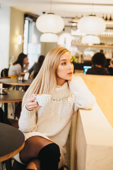 Senhora apaixonada atraente em vestido de malha segurando caneca de bebida quente e olhando para longe no café — Fotografia de Stock