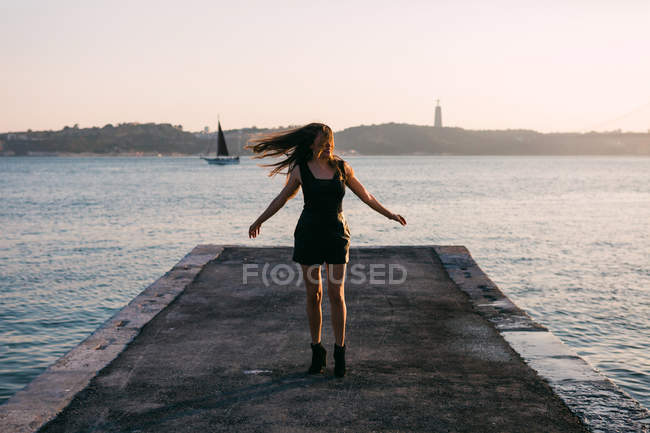 Donna allegra in abito nero e stivali che ballano su argine vicino alla superficie dell'acqua con yacht al tramonto — Foto stock