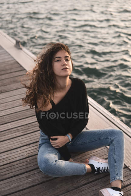 Jovem fêmea olhando para a câmera enquanto sentado no cais de madeira perto da água — Fotografia de Stock