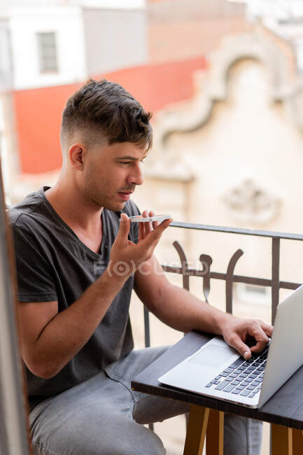 Erwachsener schöner Mann sitzt auf Balkon und schaut Laptop und sendet Sprachnachricht per Smartphone — Stockfoto
