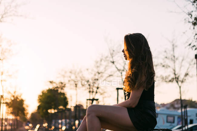 Sinnliche junge Dame in schwarzem Kleid sitzt auf Bank auf der Straße in der Gasse bei Sonnenuntergang — Stockfoto