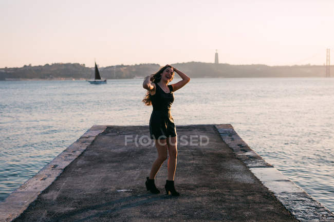 Весела жінка в чорному одязі і чоботи танцюють на набережній біля поверхні води з яхтою на заході сонця — стокове фото