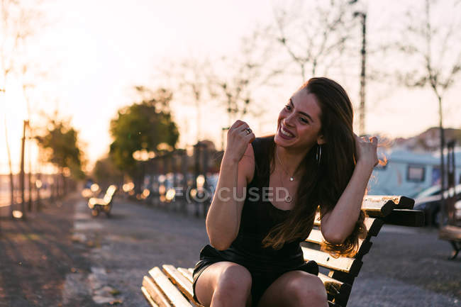 Ritratto di giovane donna allegra in abito nero seduta su panchina in strada al tramonto — Foto stock