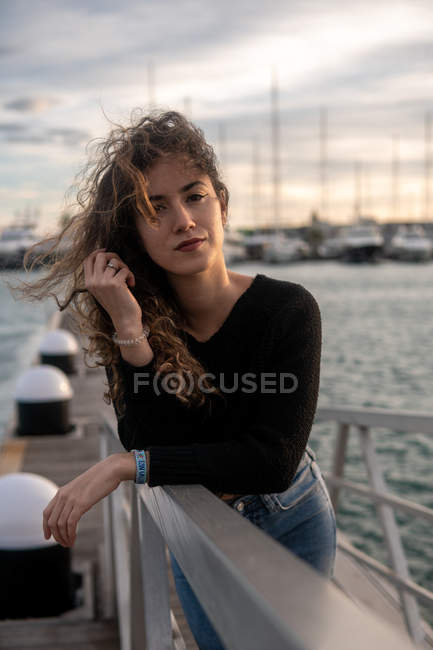 Portrait of young lady touching curly hair and looking at camera while leaning on railing on blurred background of pier and water — Stock Photo