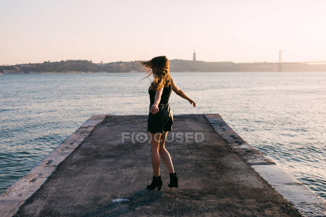 Веселая женщина в черной одежде и сапогах танцует на набережной возле поверхности воды на закате — стоковое фото