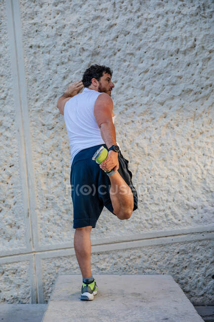 Back view of handsome adult man leaning on concrete wall and doing warming up exercise for legs during outdoor workout on street — Stock Photo
