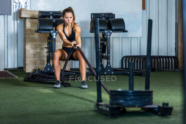Молодая женщина в спортивной одежде тренируется с боевой веревкой и конструкцией с тяжелыми пластинами в тренажерном зале — стоковое фото