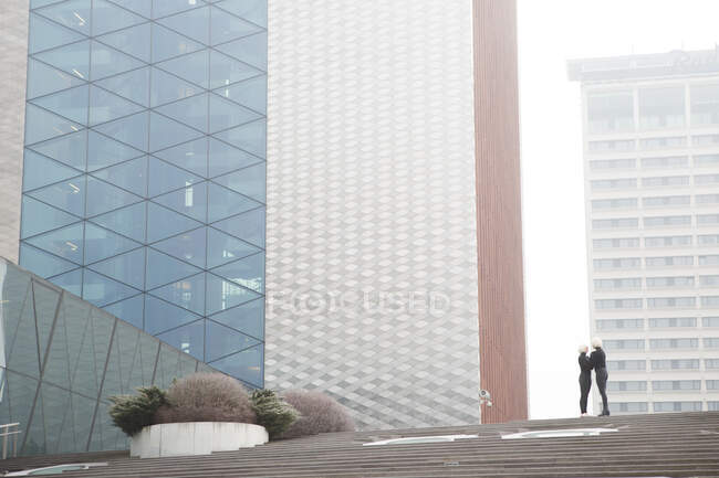 Side view women in dark wear embracing near stairs and modern building in fog in Lithuania — Stock Photo
