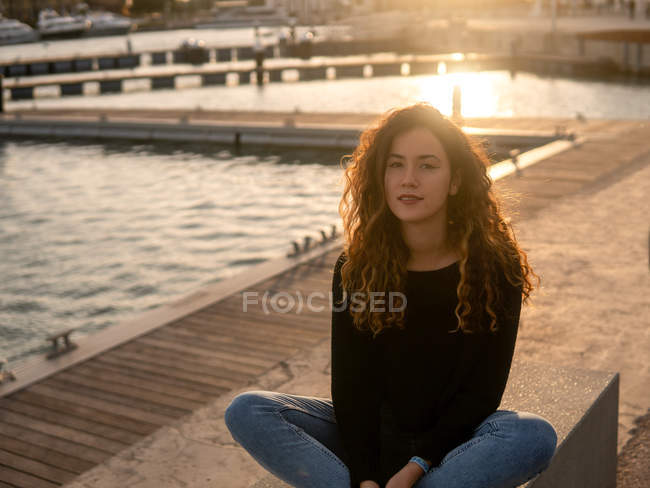Jovem mulher positiva olhando para a câmera enquanto se senta perto da água no aterro da cidade durante o pôr do sol — Fotografia de Stock