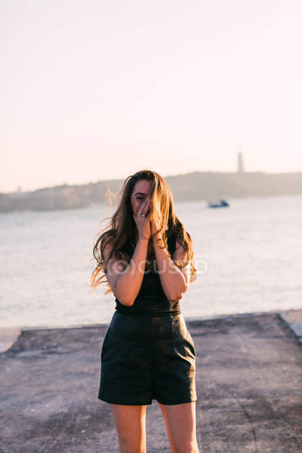 Сміється молода жінка з руками на обличчі в чорному платті, що стоїть на набережній біля поверхні води на заході сонця — стокове фото