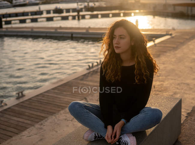 Продумана молода жінка дивиться на камеру, сидячи біля води на міській набережній під час заходу сонця — стокове фото