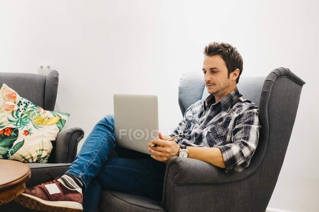 Guy in abbigliamento casual con orologio che lavora con il computer portatile e seduto alla poltrona vicino al muro bianco — Foto stock