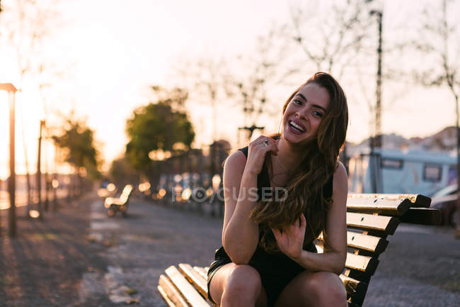 Портрет веселой молодой женщины в черном платье, сидящей на скамейке на улице на закате — стоковое фото