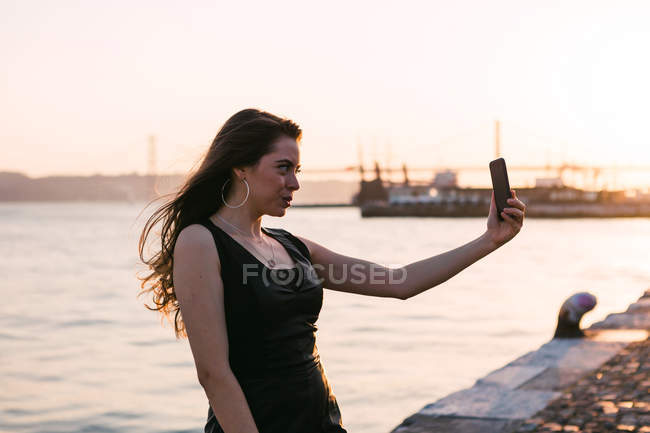 Jovem atraente em vestido preto posando e tomando selfie no dique perto da superfície da água ao pôr do sol — Fotografia de Stock