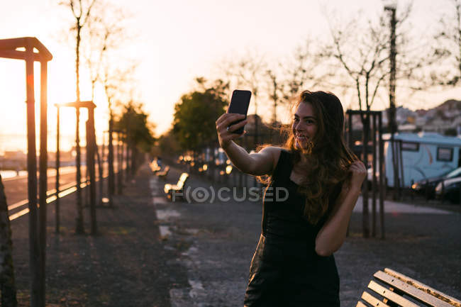 Atractiva dama en vestido negro con la mano en el pelo tomando selfie en la calle al atardecer - foto de stock