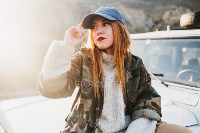 Jeune femme en tenue décontractée toucher chapeau et assis sur la voiture — Photo de stock