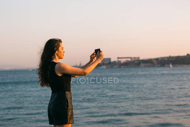 Молода жінка в чорній сукні фотографує зі смартфоном на набережній біля води на заході сонця — стокове фото
