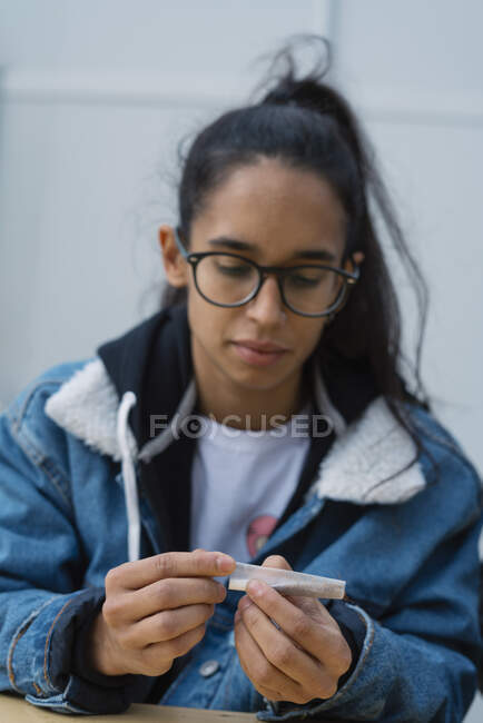 Pretty young female in casual outfit rolling cigarette while sitting on street — Stock Photo