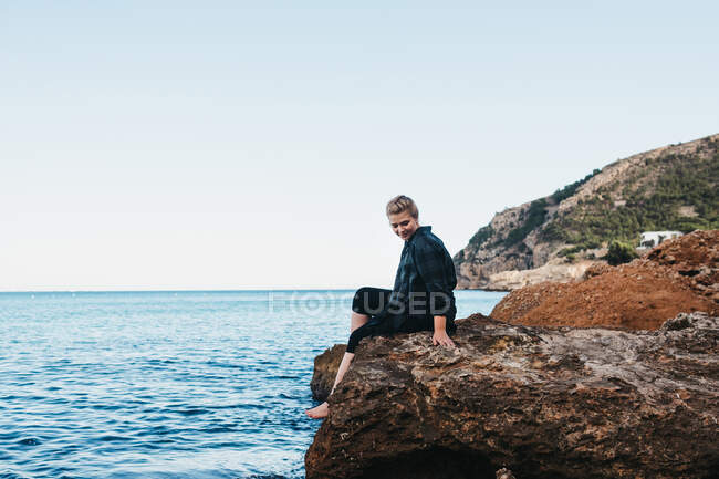 Side view of barefoot female in casual outfit sitting on stony coast near sea in Altea, Spain — Stock Photo