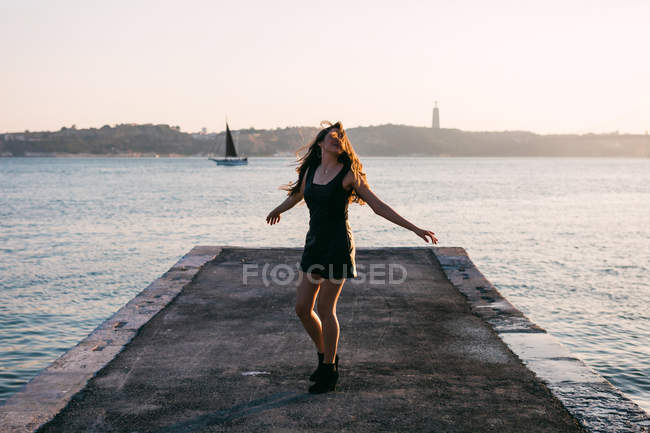 Mujer alegre en ropa negra y botas bailando en terraplén cerca de la superficie del agua con yate al atardecer - foto de stock