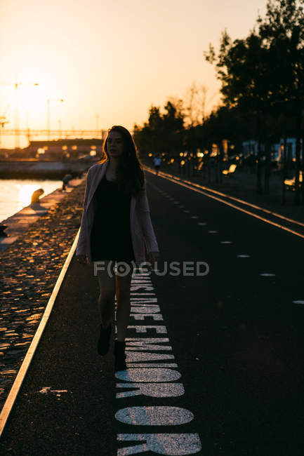 Silhouette di giovane ragazza in abito e stivali che cammina sul vicolo vicino all'acqua al tramonto — Foto stock