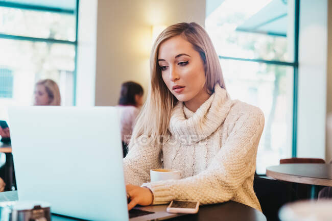 Young attractive lady in sweater typing on laptop and sitting at table near mug of coffee and mobile phone in cafe — Stock Photo